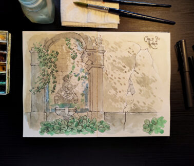 A imagem mostra uma mesa de trabalho vista de cima, com um caderno aberto exibindo uma ilustração feita em aquarela e caneta. A ilustração retrata uma estrutura antiga de pedra, semelhante a uma fonte ou nicho decorativo. No centro há uma pequena escultura de figura humana clássica, cercada por blocos de pedra irregulares e trepadeiras verdes que descem suavemente pelas laterais do arco. Na base, uma bacia de pedra e folhagens amplas em tons de verde reforçam a sensação de um jardim antigo e silencioso. O conjunto transmite uma atmosfera tranquila e contemplativa, evocando a passagem do tempo sobre a arquitetura. Ao redor do caderno, sobre a mesa escura, há materiais de pintura: à esquerda, uma paleta de aquarela usada com cores misturadas; acima, um frasco translúcido de vidro, pincéis apoiados sobre um papel toalha; à direita, três canetas de desenho. A luz é suave e quente, iluminando principalmente o centro do caderno, criando um ambiente de trabalho calmo e íntimo, como um momento de estudo artístico dedicado e sereno.
