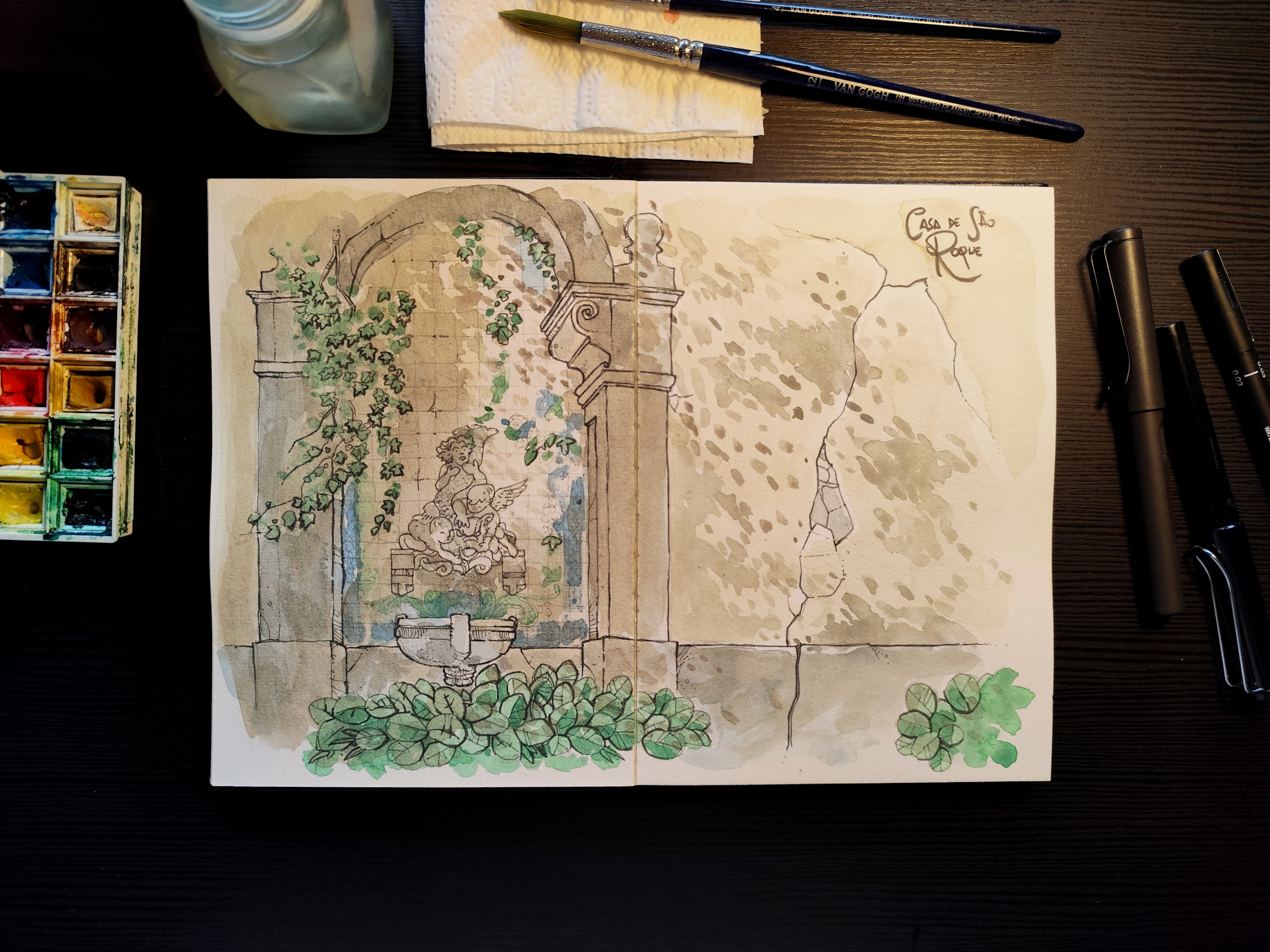 A imagem mostra uma mesa de trabalho vista de cima, com um caderno aberto exibindo uma ilustração feita em aquarela e caneta. A ilustração retrata uma estrutura antiga de pedra, semelhante a uma fonte ou nicho decorativo. No centro há uma pequena escultura de figura humana clássica, cercada por blocos de pedra irregulares e trepadeiras verdes que descem suavemente pelas laterais do arco. Na base, uma bacia de pedra e folhagens amplas em tons de verde reforçam a sensação de um jardim antigo e silencioso. O conjunto transmite uma atmosfera tranquila e contemplativa, evocando a passagem do tempo sobre a arquitetura. Ao redor do caderno, sobre a mesa escura, há materiais de pintura: à esquerda, uma paleta de aquarela usada com cores misturadas; acima, um frasco translúcido de vidro, pincéis apoiados sobre um papel toalha; à direita, três canetas de desenho. A luz é suave e quente, iluminando principalmente o centro do caderno, criando um ambiente de trabalho calmo e íntimo, como um momento de estudo artístico dedicado e sereno.