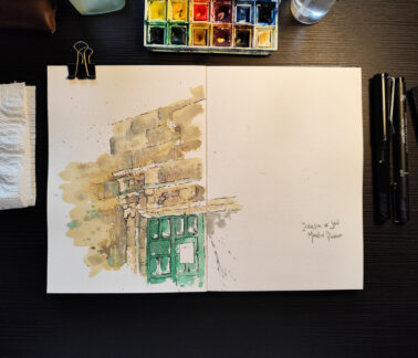 The image shows a workspace viewed from above, featuring an open, thick-paper sketchbook. On the left side of the notebook, there is an unfinished illustration, done in watercolour and pen, depicting a section of an old stone façade. The drawing shows an upper stretch of wall with irregular blocks and, below it, a doorway with architectural details. The door is green and has small rectangular windows. The watercolour uses earthy tones, soft greens, and splatters of paint, creating a light, spontaneous, and plein air study atmosphere. The right side of the page is mostly blank, save for the handwritten inscription, “Iglesia de San Martín, Dinan.” This empty area reinforces the sense of process, as if the artist is midway through the visual record of the location. Painting materials surround the sketchbook: to the left, brushes and a used paper towel; above, a watercolour palette with mixed colours and a glass jar; to the right, three drawing pens and a mechanical pencil. The setting is illuminated by soft light, creating a sense of quiet concentration and an intimate moment of creation, where the focus is on observing, interpreting, and slowly constructing the image.