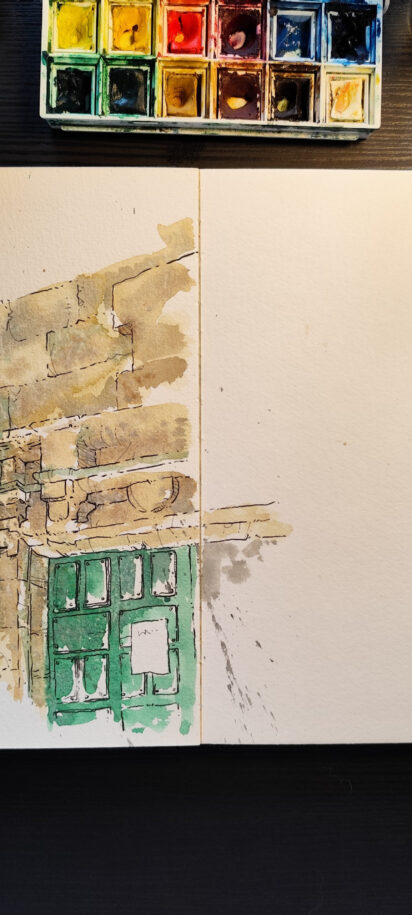 The image shows a workspace viewed from above, featuring an open, thick-paper sketchbook. On the left side of the notebook, there is an unfinished illustration, done in watercolour and pen, depicting a section of an old stone façade. The drawing shows an upper stretch of wall with irregular blocks and, below it, a doorway with architectural details. The door is green and has small rectangular windows. The watercolour uses earthy tones, soft greens, and splatters of paint, creating a light, spontaneous, and plein air study atmosphere. The right side of the page is mostly blank, save for the handwritten inscription, “Iglesia de San Martín, Dinan.” This empty area reinforces the sense of process, as if the artist is midway through the visual record of the location. Painting materials surround the sketchbook: to the left, brushes and a used paper towel; above, a watercolour palette with mixed colours and a glass jar; to the right, three drawing pens and a mechanical pencil. The setting is illuminated by soft light, creating a sense of quiet concentration and an intimate moment of creation, where the focus is on observing, interpreting, and slowly constructing the image.