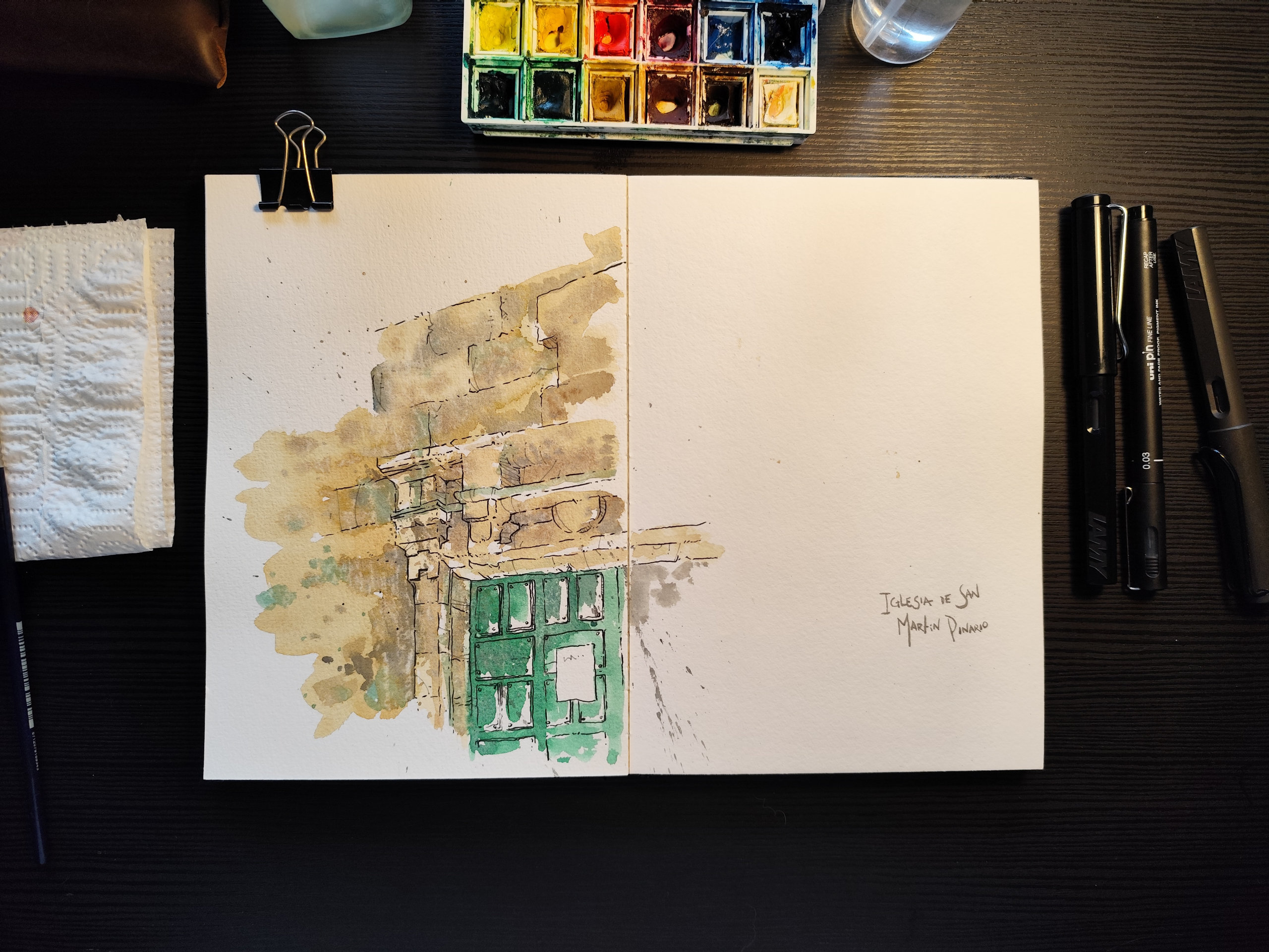 The image shows a workspace viewed from above, featuring an open, thick-paper sketchbook. On the left side of the notebook, there is an unfinished illustration, done in watercolour and pen, depicting a section of an old stone façade. The drawing shows an upper stretch of wall with irregular blocks and, below it, a doorway with architectural details. The door is green and has small rectangular windows. The watercolour uses earthy tones, soft greens, and splatters of paint, creating a light, spontaneous, and plein air study atmosphere. The right side of the page is mostly blank, save for the handwritten inscription, “Iglesia de San Martín, Dinan.” This empty area reinforces the sense of process, as if the artist is midway through the visual record of the location. Painting materials surround the sketchbook: to the left, brushes and a used paper towel; above, a watercolour palette with mixed colours and a glass jar; to the right, three drawing pens and a mechanical pencil. The setting is illuminated by soft light, creating a sense of quiet concentration and an intimate moment of creation, where the focus is on observing, interpreting, and slowly constructing the image.